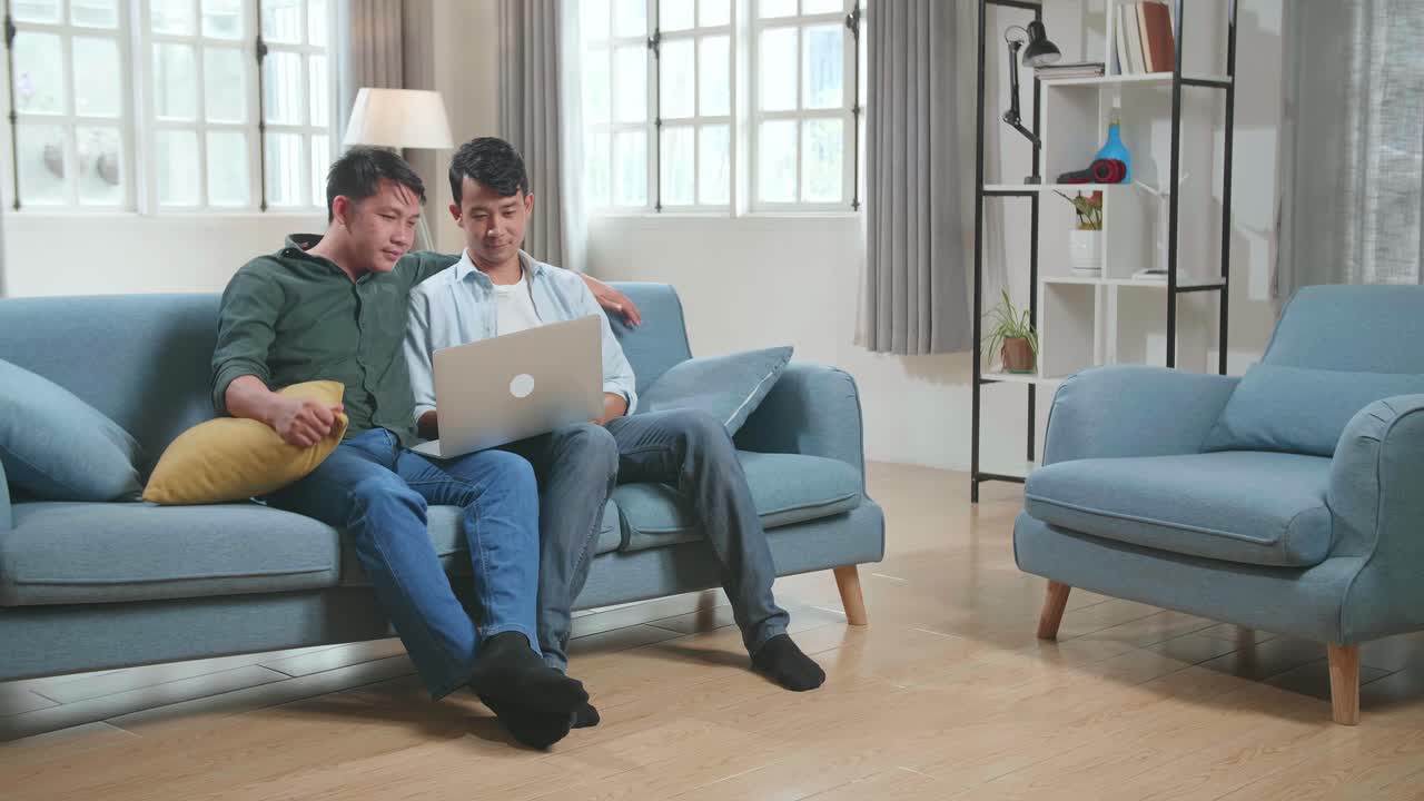 Portrait Of Asian Gentle Gay Couple Using Laptop Computer, While Sitting On A Couch In Cozy Stylish Apartment. Males Looking Each Other