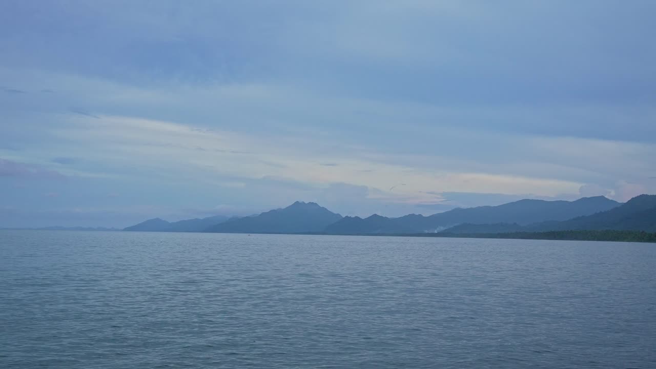 A handheld shot of the gentle sea water ripples shimmer under soft overcast light, mountains fading in the distance of a serene, layered coastal composition of Mauban Port, Quezon Province Philippines