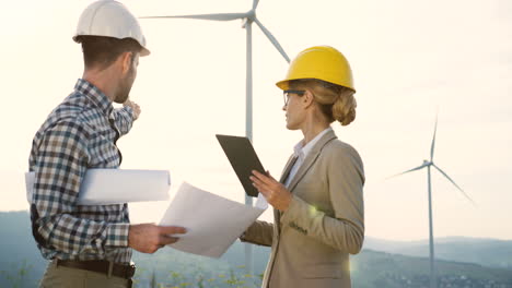 caucasian man and woman engineers wearing a helmet watching some blueprints and using tablet while talking at wind station of renewable energy