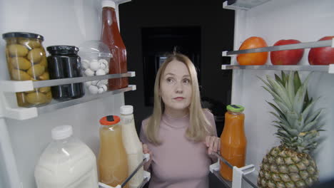 a woman looks inside a large refrigerator and drinks milk from a glass. snack on the go