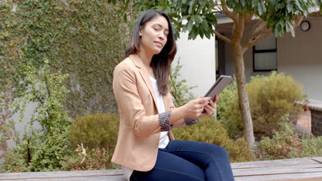 young biracial woman sits on a bench outdoors, holding a tablet