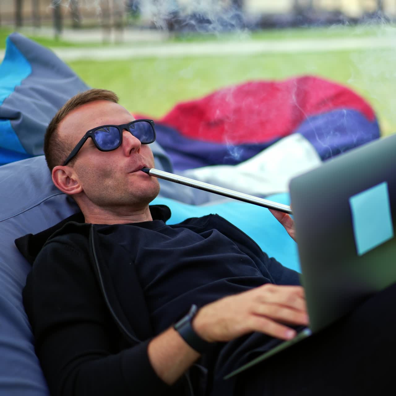 Smiling male freelancer working on computer combining it with rest outdoors. Man smoking hookah producing thick smoke