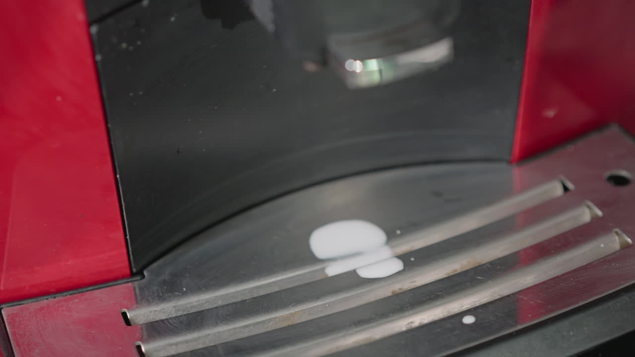 Close-up view of espresso machine nozzle dripping milk onto a stainless steel tray. The milk is spilling in small droplets, highlighting the process of steaming or foaming milk for a coffee drink