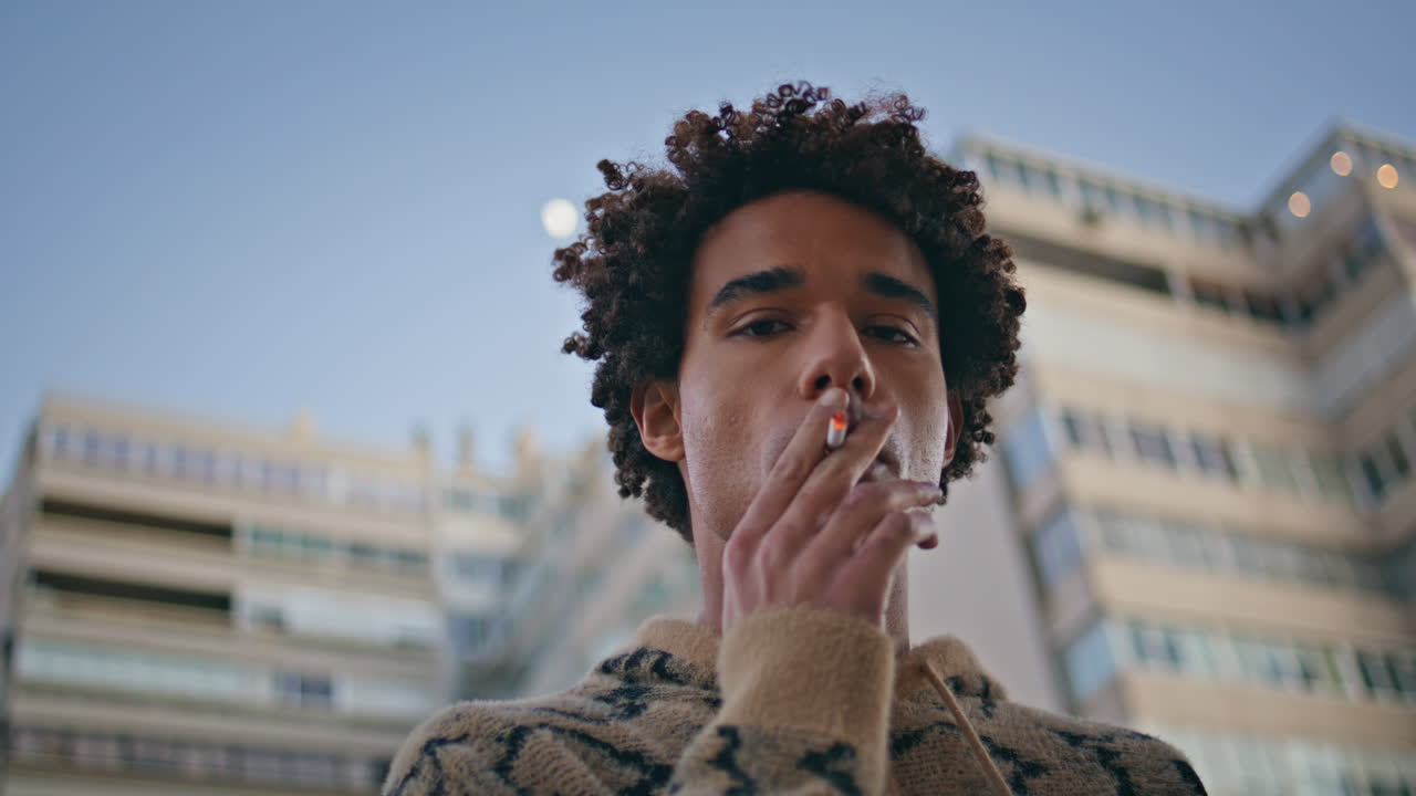 un tipo de primer plano fumando pensativamente en la ciudad. un joven hipster de cabello rizado adicto