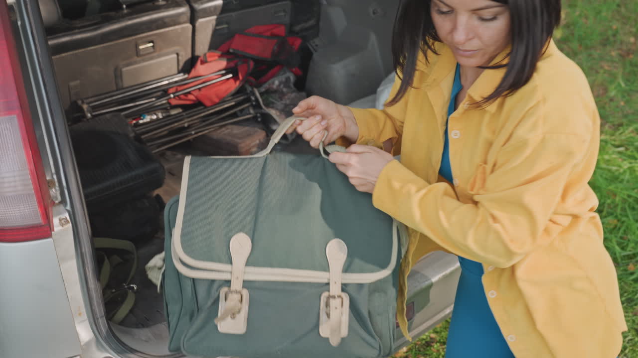 Female camper organizing gear. Woman arranging backpack straps on grass for camping activity. Female camper diligently securing and organizing supplies from car trunk for outdoor adventure