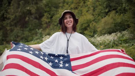 portrait of a brunette girl in a white t-shirt who stands with the flag of the united states of america that flutters in the wind against the background of a green forest