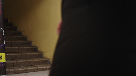 woman in grey beanie and puffer jacket sipping drink from green cup while holding leash of whippet in purple coat, standing indoors near stairs and window during cold day, calm relaxed winter moment