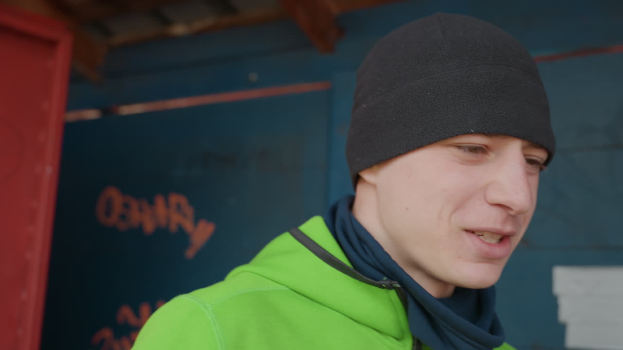 Giovane uomo bianco con felpa verde appoggiato a un muro di graffiti, che sistema la manica e la cerniera, con berretto e scaldacollo visibili, sorriso rilassato, atmosfera urbana spontanea, abbigliamento a strati per clima fresco, sfondo materico