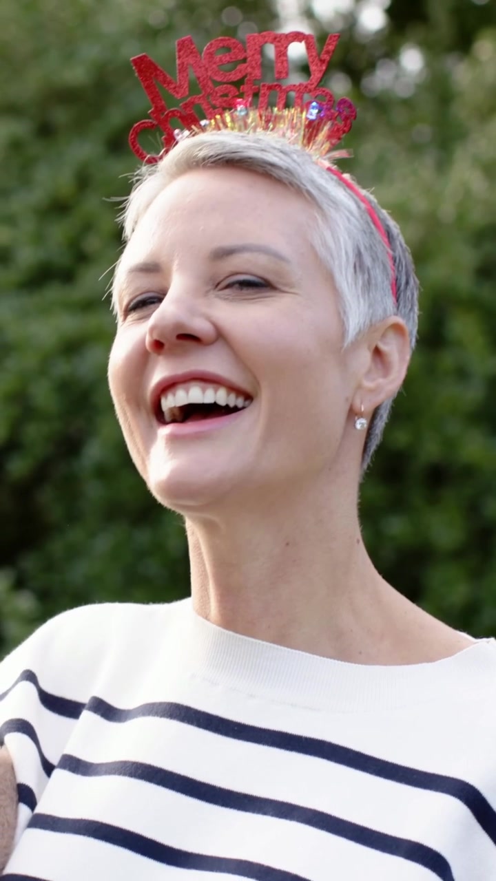 Video verticale: Donna sorridente in primo piano che si volta a destra, ridendo in giardino con una fascia 'Merry Xmas'.