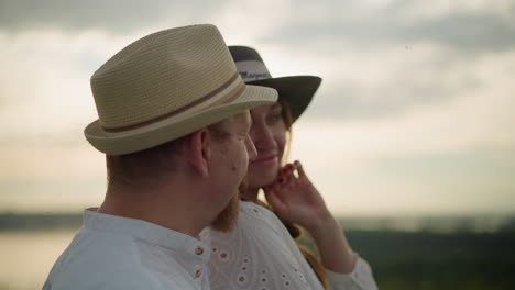 a close-up of two lovers, both wearing white shirts and hats, standing together at sunset. the man gently holds the woman from behind, capturing a moment of intimacy and warmth as the golden light