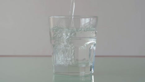 Gießen-Von-Wasser-In-Glas-Auf-Dem-Tisch-Mit-Reflektion,-Immer-Noch-Extreme-Nahaufnahme-In-Minimalistischer-Einrichtung