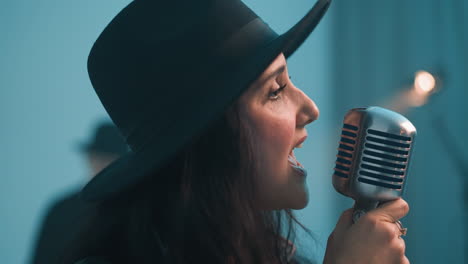 cantante apasionado con un sombrero negro y una chaqueta de cuero cantando en un micrófono vintage contra un fondo azul. otra persona está borrosa en el fondo