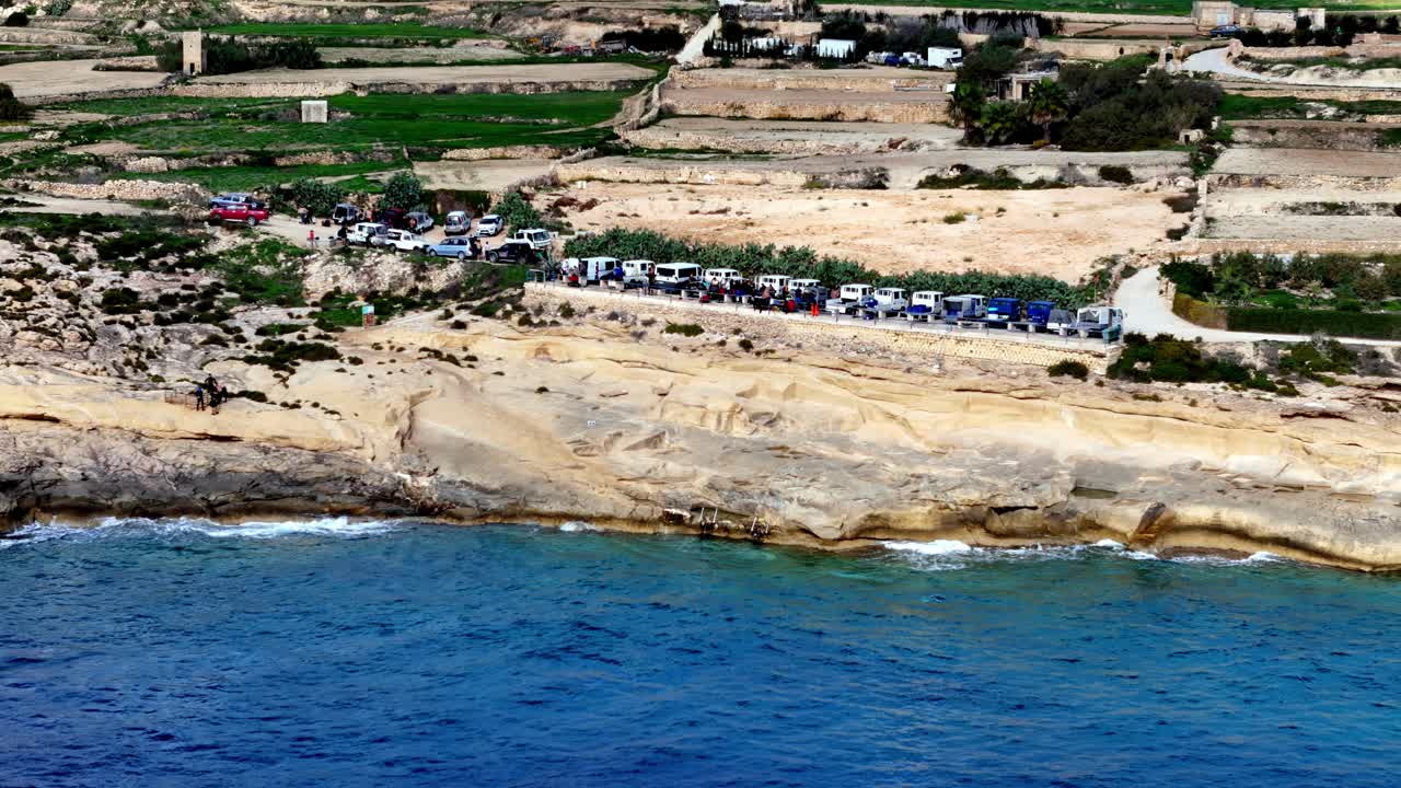 Vista aerea di camion da immersione parcheggiati sulla costa calcarea della baia di Xwejni, Gozo, Malta, durante una soleggiata mattinata primaverile con mare Mediterraneo blu intenso e campi agricoli