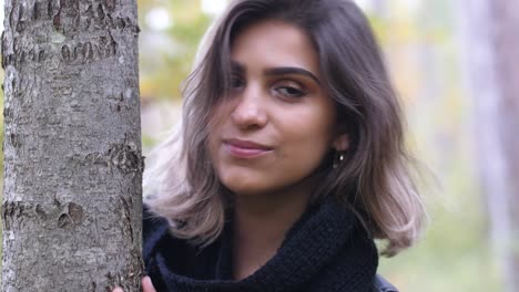 brunette girl in the autumn forest stands by a tree and smiles at the camera - close shot