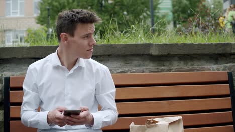 man sitting on bench with packagings with fast food, using phone with smile