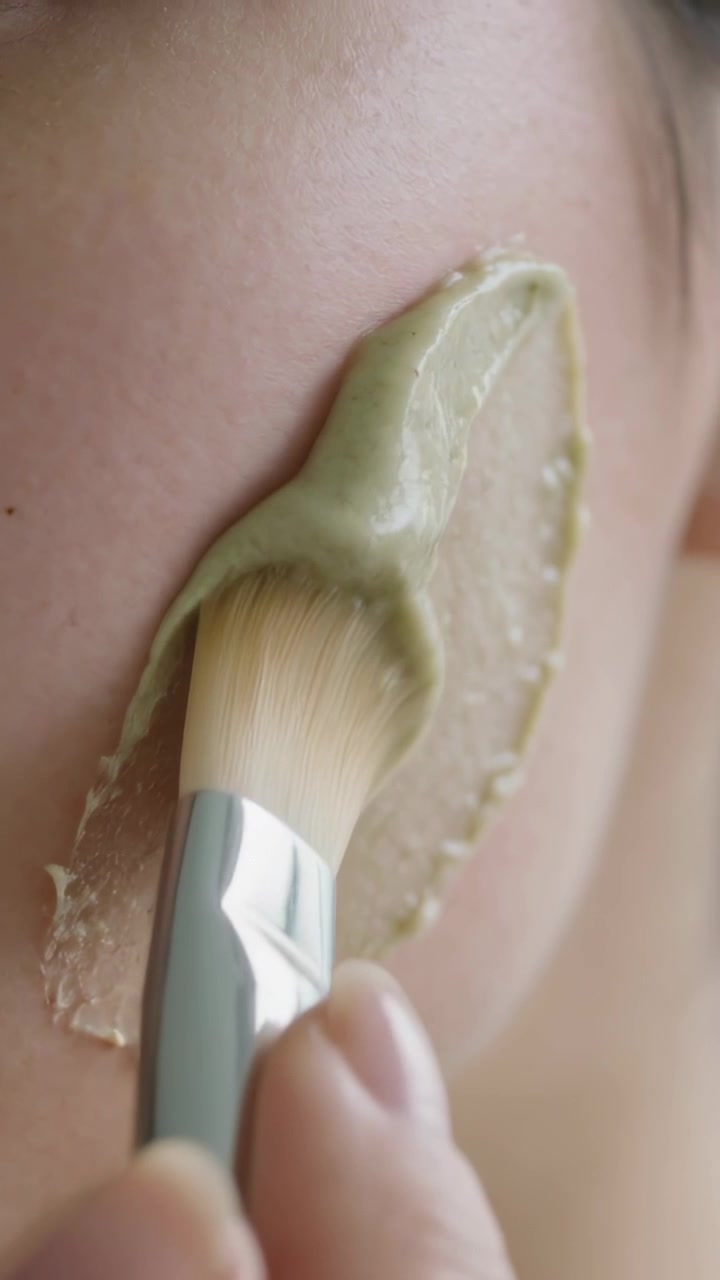 Video verticale: Spennellare la guancia, con il pennello che preleva una noce di prodotto dal lavandino, applicando una maschera verde per una pelle più uniforme.