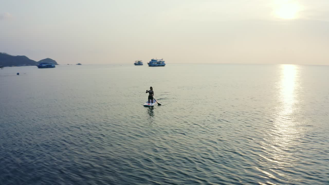 A picturesque aerial shot of a woman paddle boarding in the calm waters of Koh Tao during golden hour.