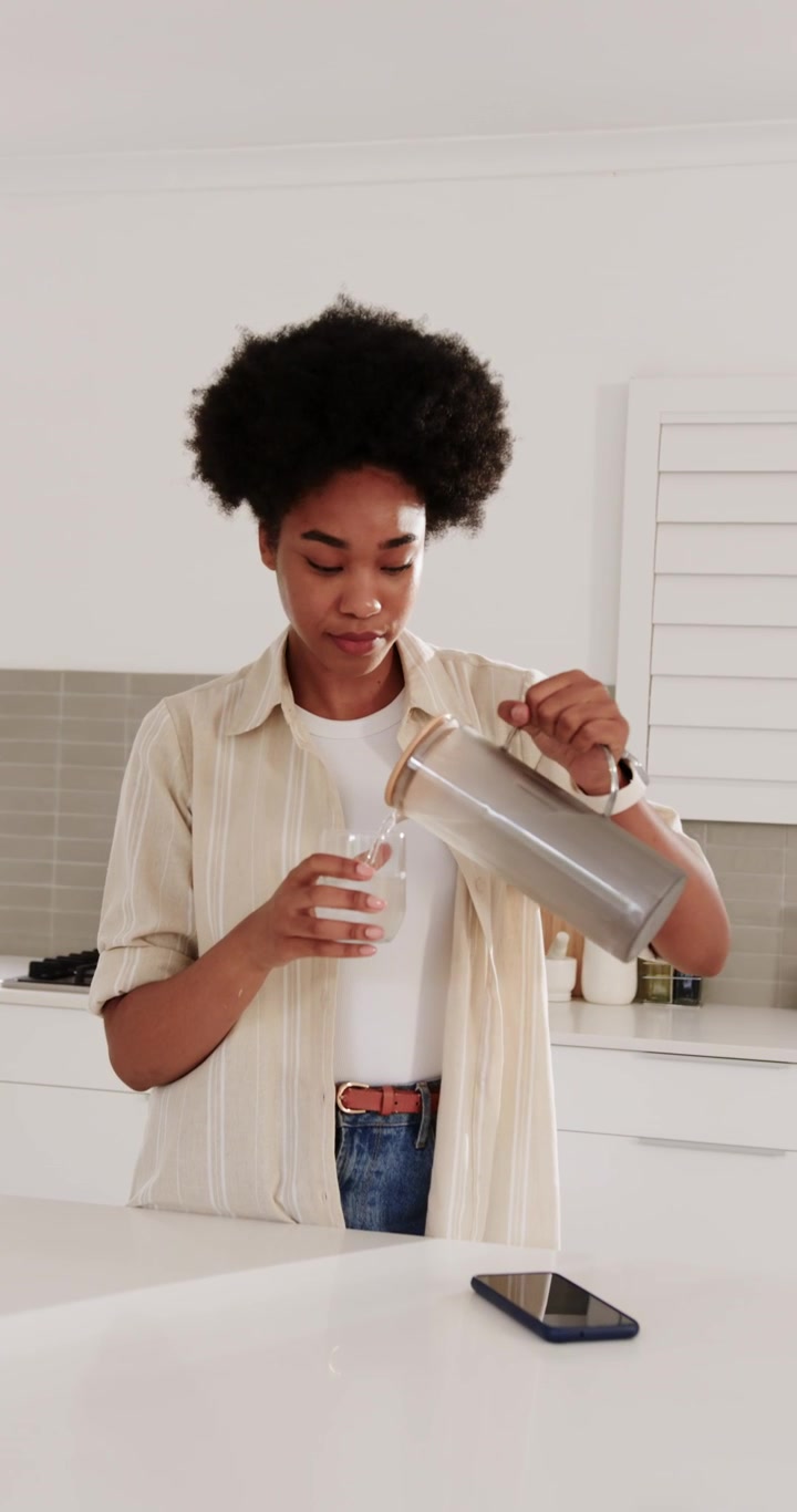 Video verticale: Donna che, sull'isola della cucina, inclina una caraffa, versa un bicchiere, sorseggia e la sposta con il telefono.