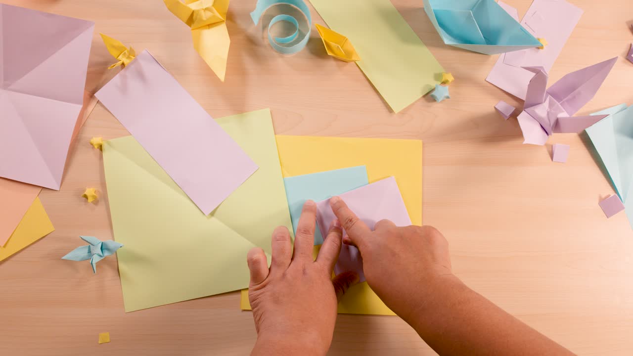 Person folds pastel origami paper on wooden table, surrounded by finished models, under soft lighting