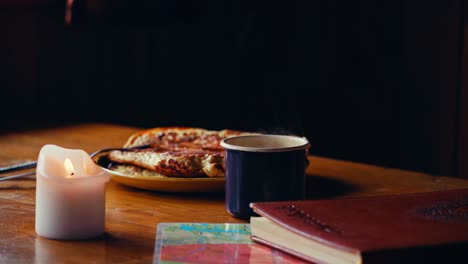 wooden table with lit candle next to steaming mug of coffee, plate of pancakes, and books in foreground. static shot