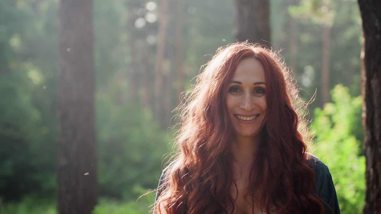 Portrait of female parent with long red hair smiling warmly while standing in lush forest during sunny day, soft natural light illuminating her face, cheerful atmosphere of peaceful outdoor moment