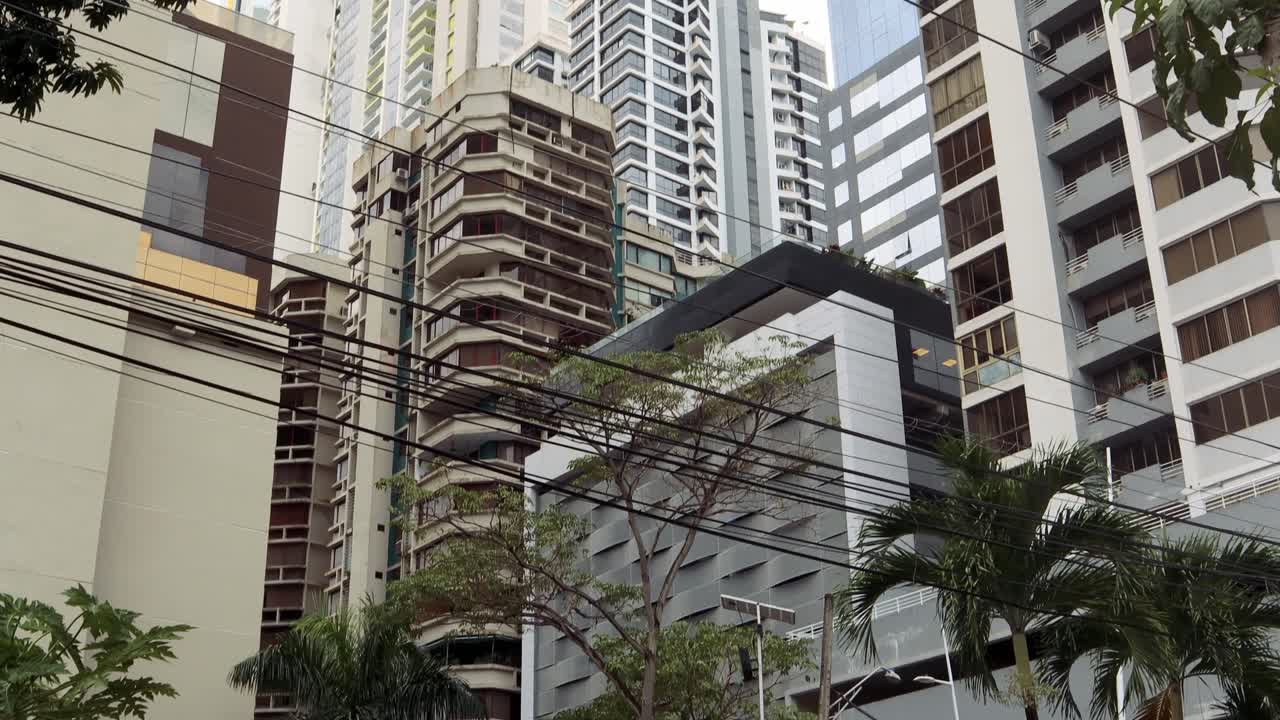 4K descending view of a cluster of tall, close-knit modern buildings on a famous avenue with tropical vegetation in Panama City