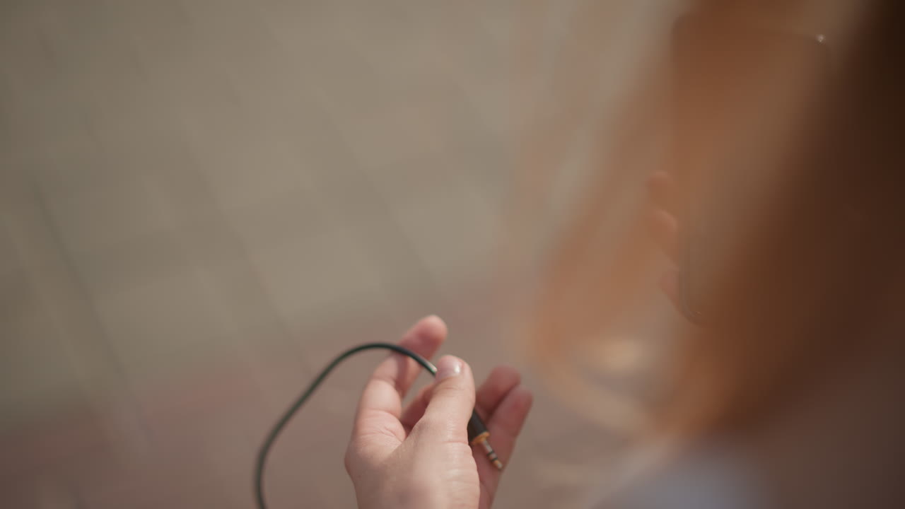 blonde woman plugging earphones into smartphone on sunlit street, hands connect cord to device, tapping screen to select playlist. Casual urban atmosphere suggests dancer preparing for street