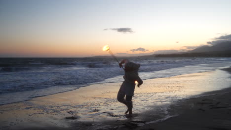 un hombre bailarín de fuego en silueta girando su bastón en llamas en la playa al atardecer con llamas y olas del océano a cámara lenta