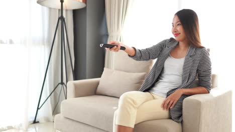 Joven-Mujer-Asiática-Adulta-Elegante-Ropa-Casual-Sentada-En-El-Sofá-Con-Control-Remoto-En-La-Mano-Zapping,-Viendo-Televisión-Y-Saltando-De-Canal-De-Televisión