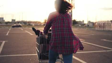 vista trasera de una mujer joven empujando el carrito de la compra, mientras su amiga está sentada adentro durante la puesta de sol