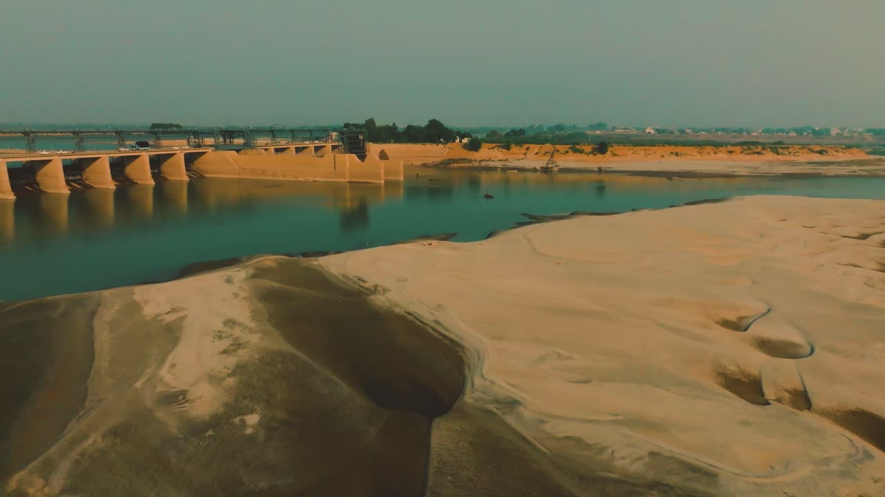 Banco di sabbia, acqua del fiume e diga nel Sindh, Pakistan. Il traffico transita sul ponte sotto un intenso cielo pomeridiano velato, arancione e verde acqua. Panoramica aerea a sinistra in 4K.