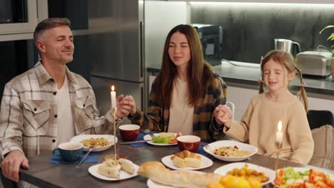 Familia-Feliz,-Mujer-Morena-Con-Una-Camisa-A-Cuadros-Junto-Con-Su-Esposo-De-Mediana-Edad-Con-Cabello-Gris-Y-Su-Pequeña-Hija-Rezando-Y-Dando-Gracias-Por-La-Comida-Antes-De-Comenzar-Una-Cena-En-Una-Cocina-Moderna