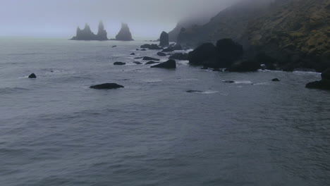 Aerial-cinematic-slow-pan-up-waves-crashing-on-haystacks-shoreline-at-Vik-Iceland-early-winter-fog-at-Black-Sand-Beach
