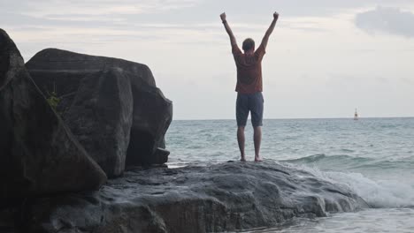 Asiatischer-Mann-Hebt-Die-Arme,-Während-Er-Bei-Flut-Auf-Dem-Felsen-Mit-Blick-Auf-Das-Meer-Am-Strand-Von-Dam-Trau-In-Vietnam-Steht