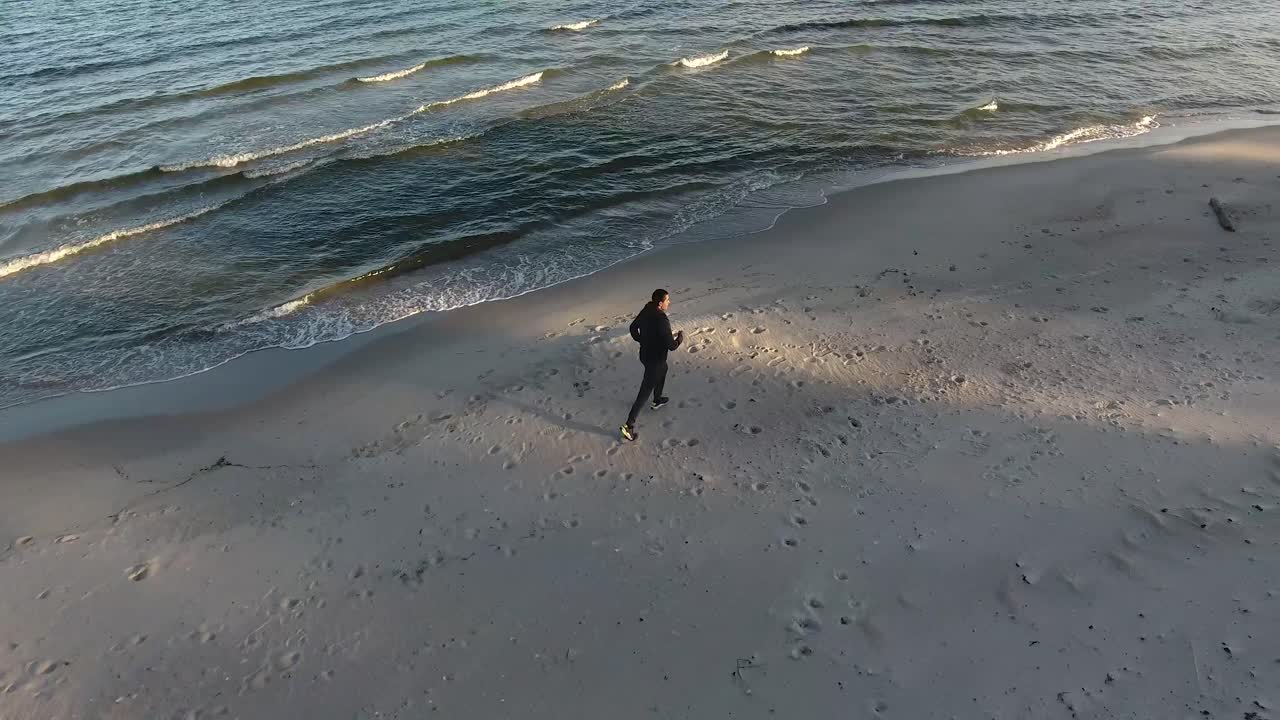 toma aérea de seguimiento de un hombre corriendo junto a las olas en la playa al atardecer en ystad, sur de suecia, skåne en el verano