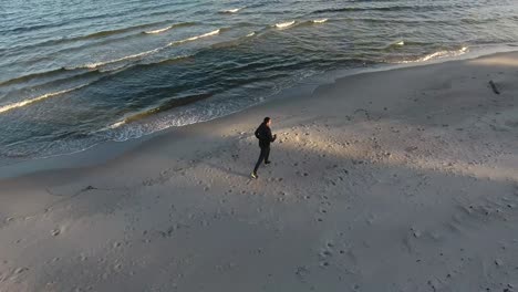 Toma-Aérea-De-Un-Hombre-Corriendo-Por-Las-Olas-En-La-Playa-Al-Atardecer-En-Ystad-Sur-De-Suecia-Skåne-En-El-Verano