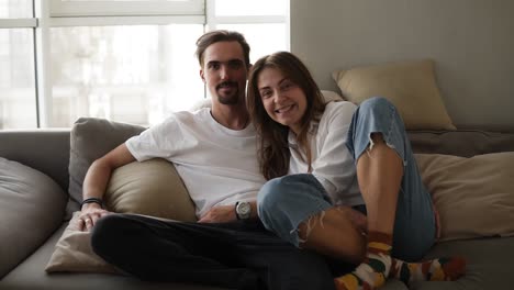 portrait shot of the young caucasian smiled bearded boyfriend and girlfriend sitting on the grey sofa and posing to the camera