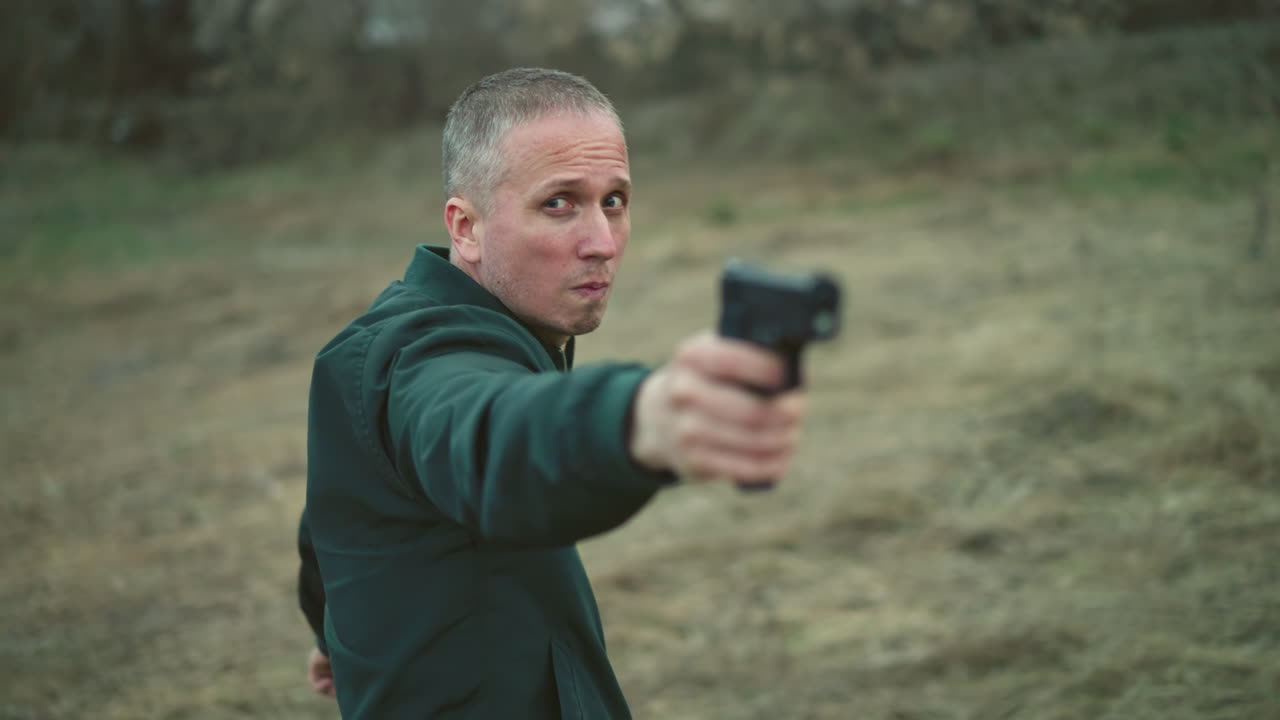 una vista cercana de un hombre con una chaqueta verde apuntando a una pistola mientras se da la vuelta en un entorno al aire libre cubierto de hierba