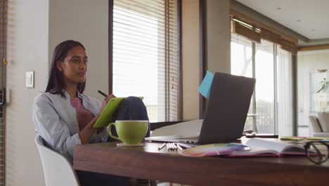 mujer de raza mixta usando una computadora portátil escribiendo en un cuaderno bebiendo café trabajando desde casa