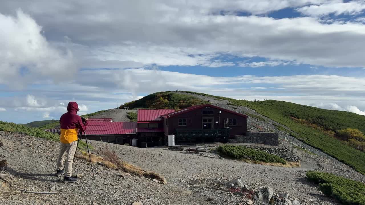 Wide shot of a hiker using a camera on a tripod to take pictures on a rocky slope facing Chogatake Hut under a bright sky