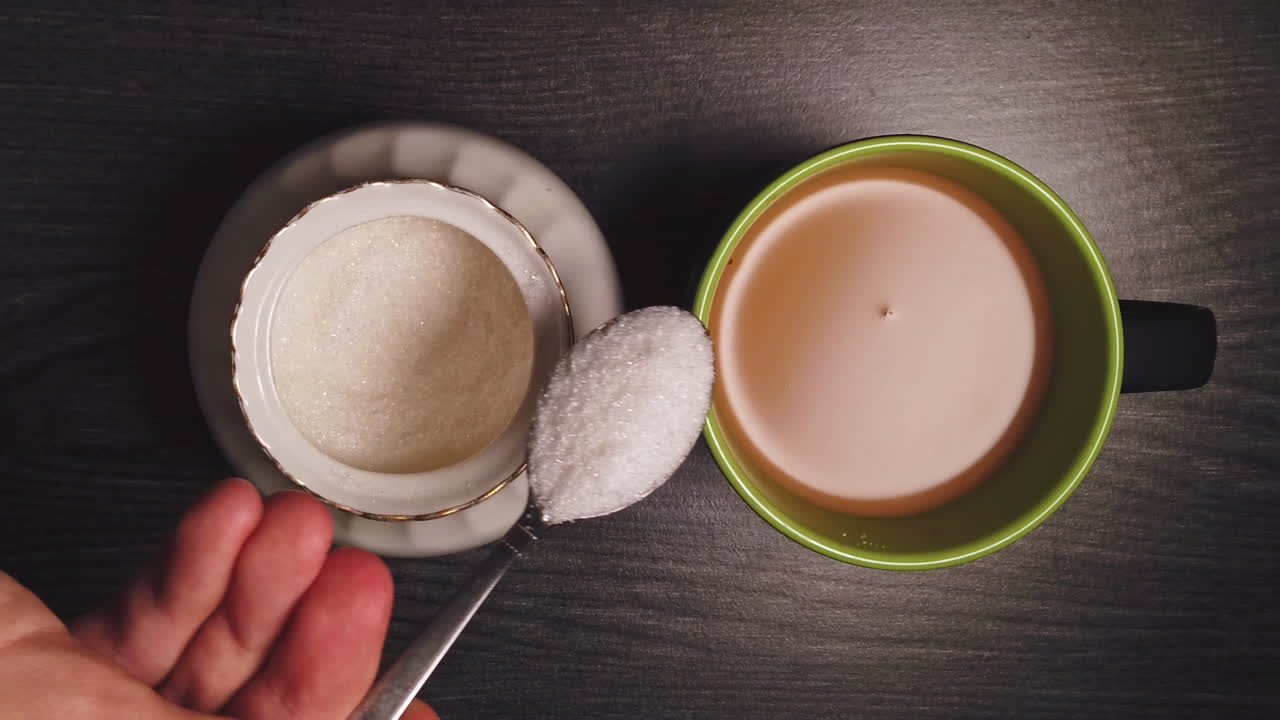 Overhead slow motion of pouring sugar from a bowl in the ceramic cup with black coffee with milk by a caucasian man on wooden table