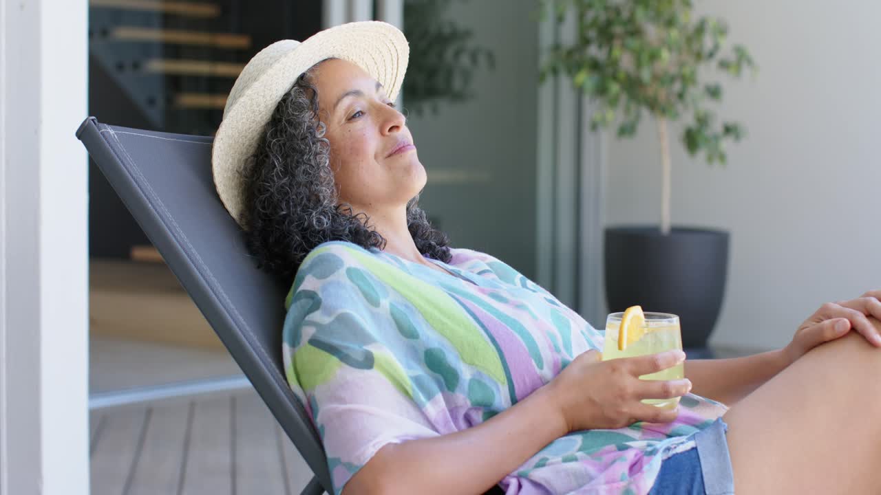 Afroamerikansk kvinde liggende på en solrig terrasse, iført stråhat, nipper til en citrusdrik og slapper af