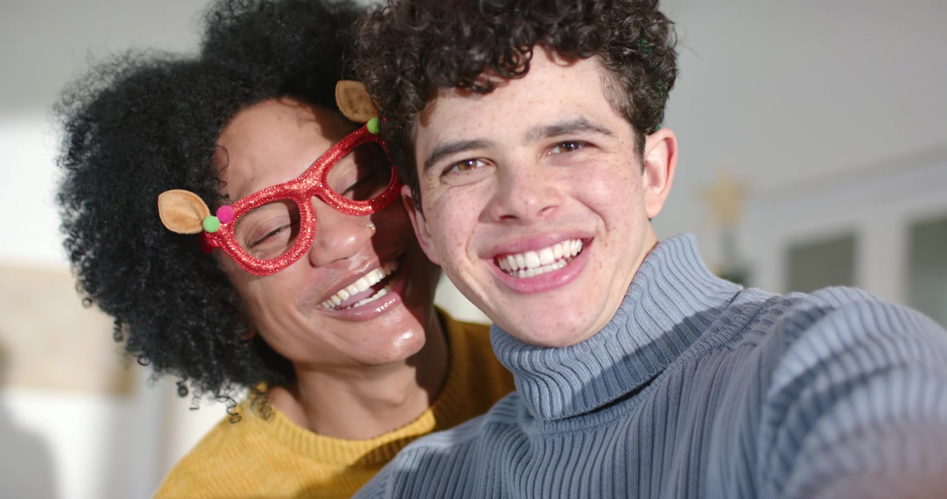 Amici di diverse etnie che si scattano un selfie a casa, quello a destra tiene il telefono, quello a sinistra indossa occhiali rossi, per divertimento.