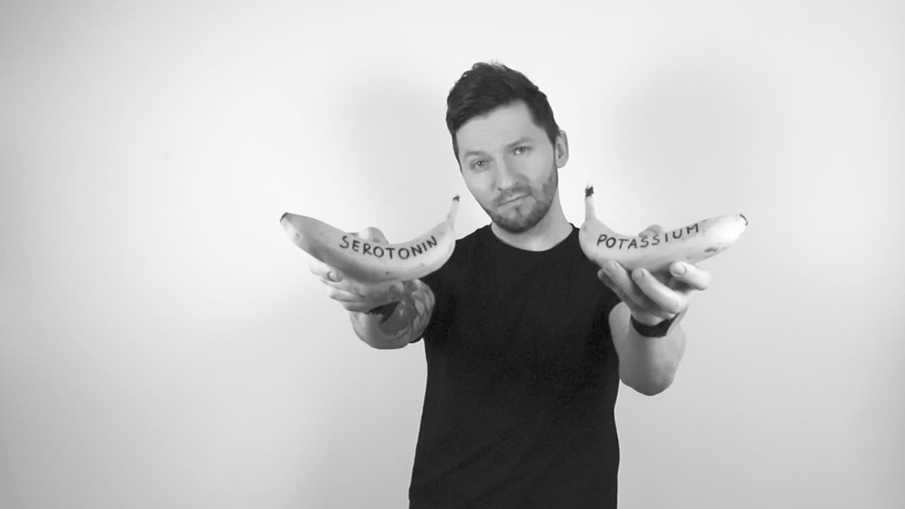 Grayscale footage of a tattooed man with short hair and trimmed beard lifting up two bananas in his hand with writing on them that says Potassium and other one says Serotonin, man is happy and smiles.