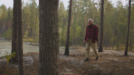 Un-Excursionista-Solitario-Y-Anciano-Camina-Por-El-Bosque-En-La-Orilla-Alta-Del-Embalse-Sosteniendo-Una-Caña-En-La-Mano-Mochilero-Y-Pescando-Con-Caña