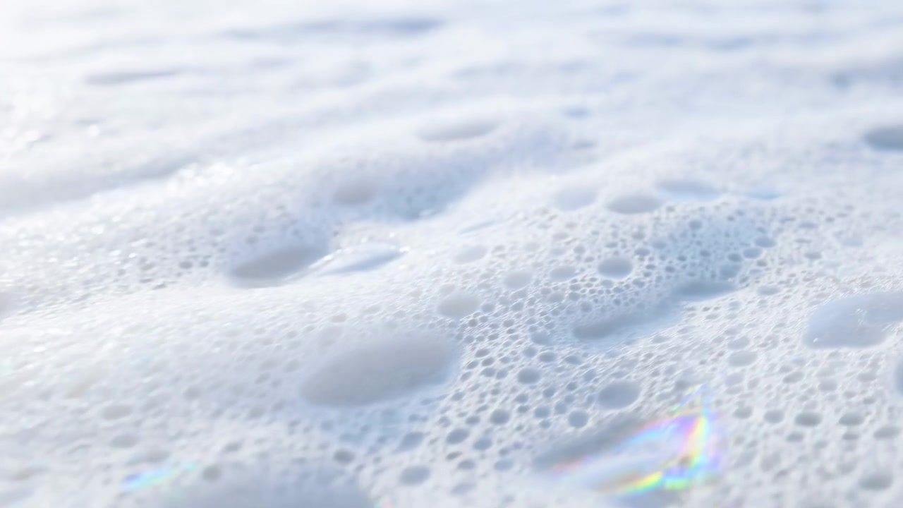 Morbida schiuma bianca con piccole bolle e leggere increspature riempie l'inquadratura, creando una superficie astratta e pulita con un aspetto fresco e calmo e una sottile luce naturale.