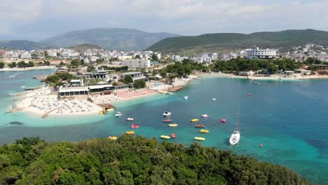 Drone-view-in-Albania-flying-over-white-sand-beaches,-blue-crystal-clear-water,-green-small-islands,-boats-and-hotels,-sunny-day-in-Ksamil