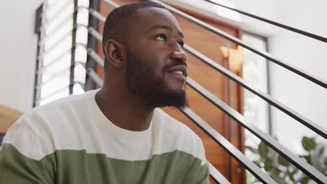 video of happy african american man smiling, sitting on stairs and looking around at home