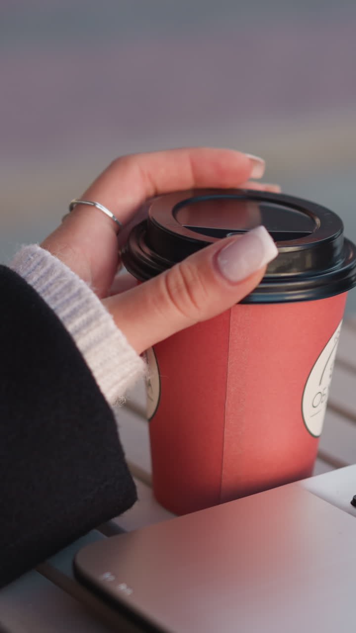 primer plano de la mano de la dama girando suavemente la taza de café con los dedos cuidados, la computadora portátil borrosa en el fondo creando un ambiente de trabajo sofisticado