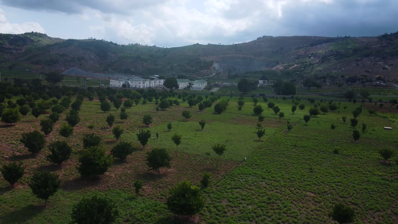 Aerial of hilly farmland landscape with crops, estate housing, scattered trees and reddish soil in Nigeria. Seamless drone view of vegetation across hill top in new develop urban area, Abuja, Africa